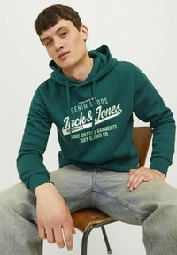 Grøn hættejakke lavet af bomuld med et grafisk print i hvid. Har snoretræk i hætten og ribbede manchetter. Båret med lyseblå jeans.