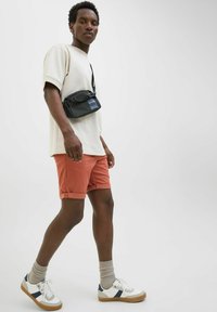Black crossbody bag, beige oversized t-shirt, rust-colored shorts, grey socks, and white sneakers with navy accents. Plain background.
