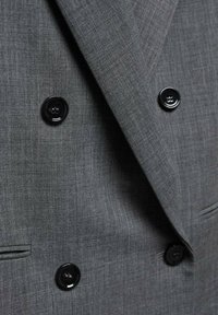 Gros plan sur un blazer croisé gris avec des boutons noirs à quatre trous et une poche fendue.