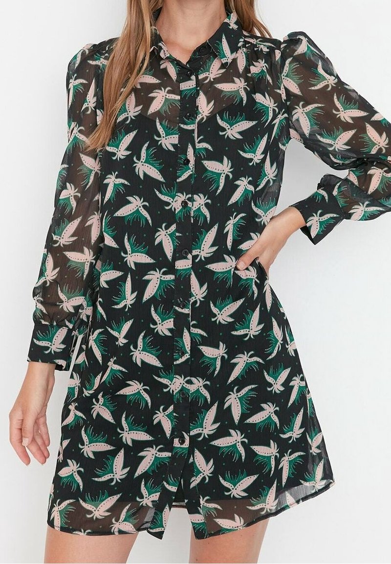 Femme portant une robe noire transparente à boutons avec des manches longues bouffantes et un motif d'oiseaux verts et roses.