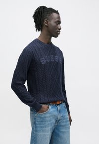 Mann mit Dreadlocks steht seitlich, trägt einen marineblauen, kabelgestrickten Pullover mit der Aufschrift "GUESS" und hellblaue Jeans mit braunem Gürtel.