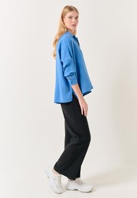 Chemise oversized bleue à manches longues, avec poignets boutonnés et fentes sur les côtés, associée à un pantalon large noir et des baskets blanches.