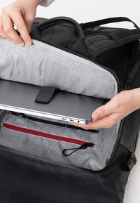 Zaino nero con interno grigio imbottito, dotato di scomparto per laptop fissato da una cinghia e una tasca rossa con zip per accessori.