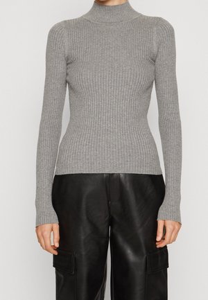 Personne portant un pull col roulé côtelé gris ajusté et un pantalon cargo en cuir noir ample, debout devant un fond uni.