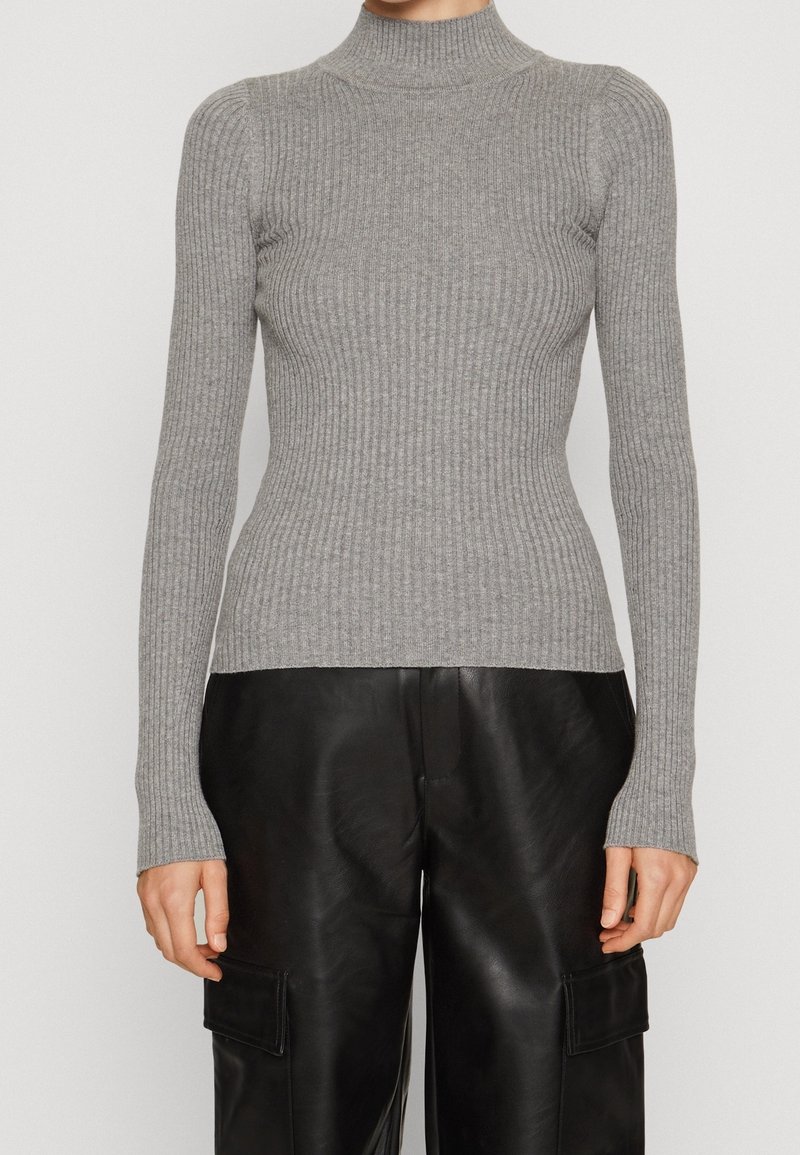 Personne portant un pull col roulé côtelé gris ajusté et un pantalon cargo en cuir noir ample, debout devant un fond uni.