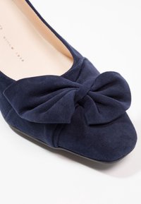 Ballerine en daim bleu marine avec un bout arrondi et un grand nœud sur le devant, dotée d'une doublure intérieure lisse et d'une semelle plate.