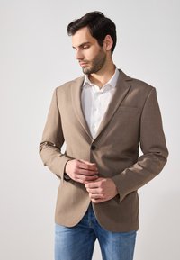 Brown blazer with a smooth fabric, single button closure, and notch lapels. Worn over a white shirt, paired with light blue jeans.