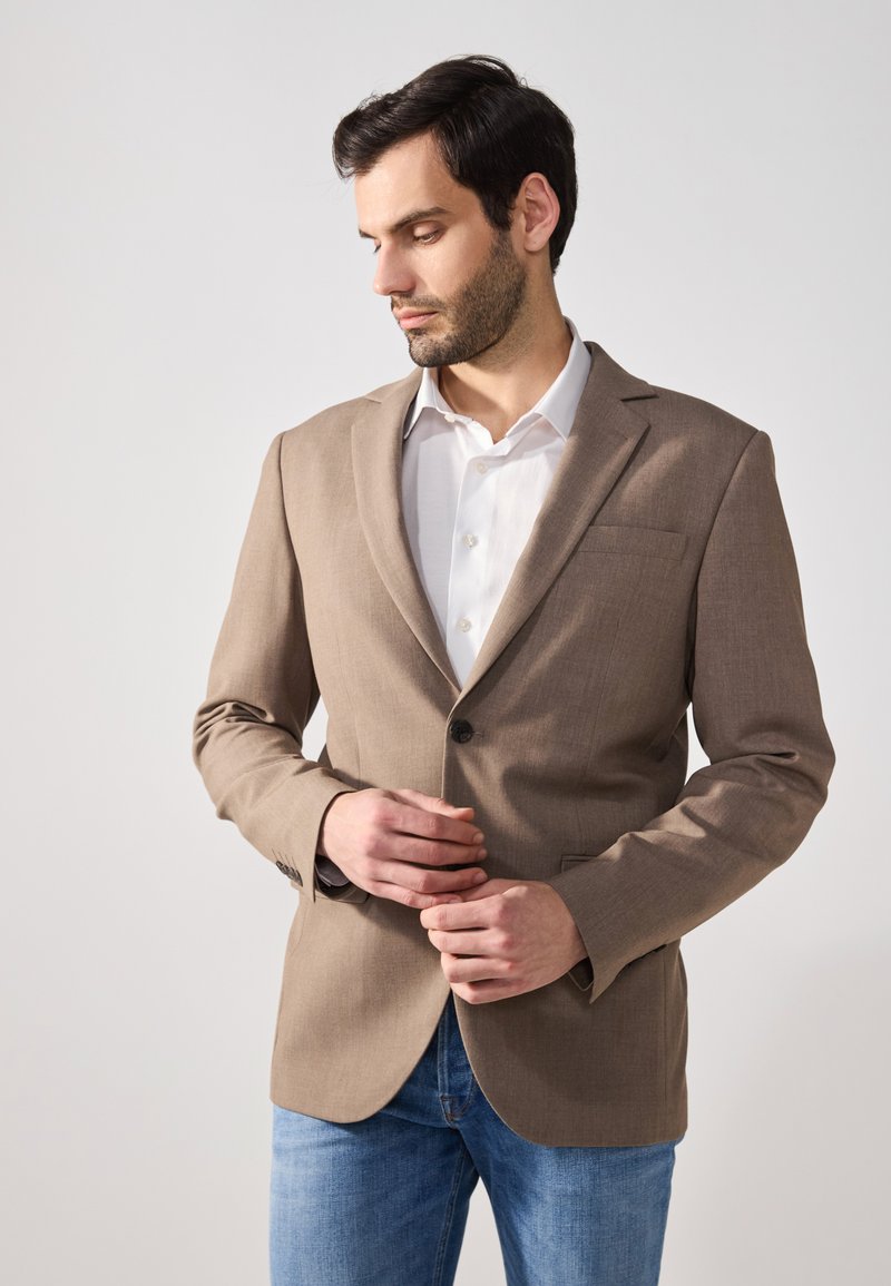 Brown blazer with a smooth fabric, single button closure, and notch lapels. Worn over a white shirt, paired with light blue jeans.