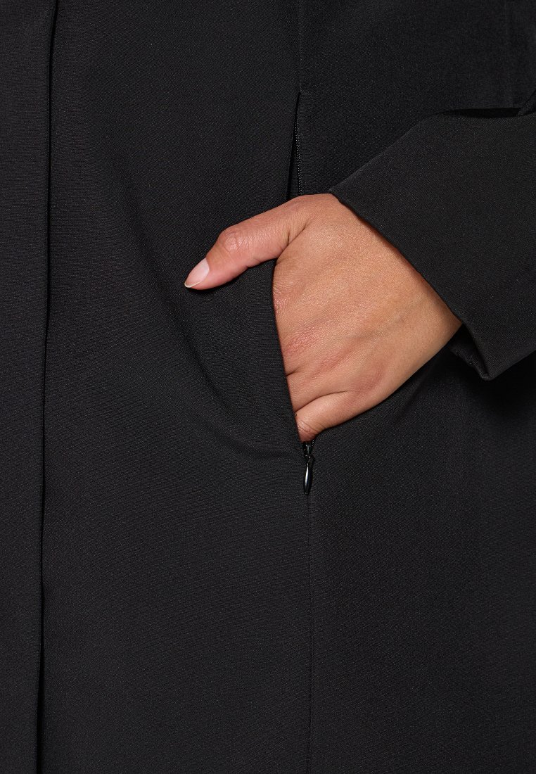 Czarna odzież o gładkiej fakturze, z boczną kieszenią i widocznym zamkiem błyskawicznym. Ręka umieszczona na kieszeni dla szczegółów.