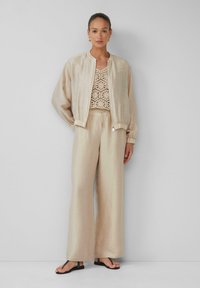Beige bomberjack met elastische manchetten, gecombineerd met een beige gehaakte top en losse broek, afgemaakt met zwarte teenslippers.