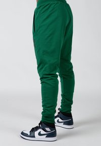 Groene joggers van zacht materiaal, met een getailleerd ontwerp, zijzakken en een donker logo-detail. Gecombineerd met zwart-witte sneakers.