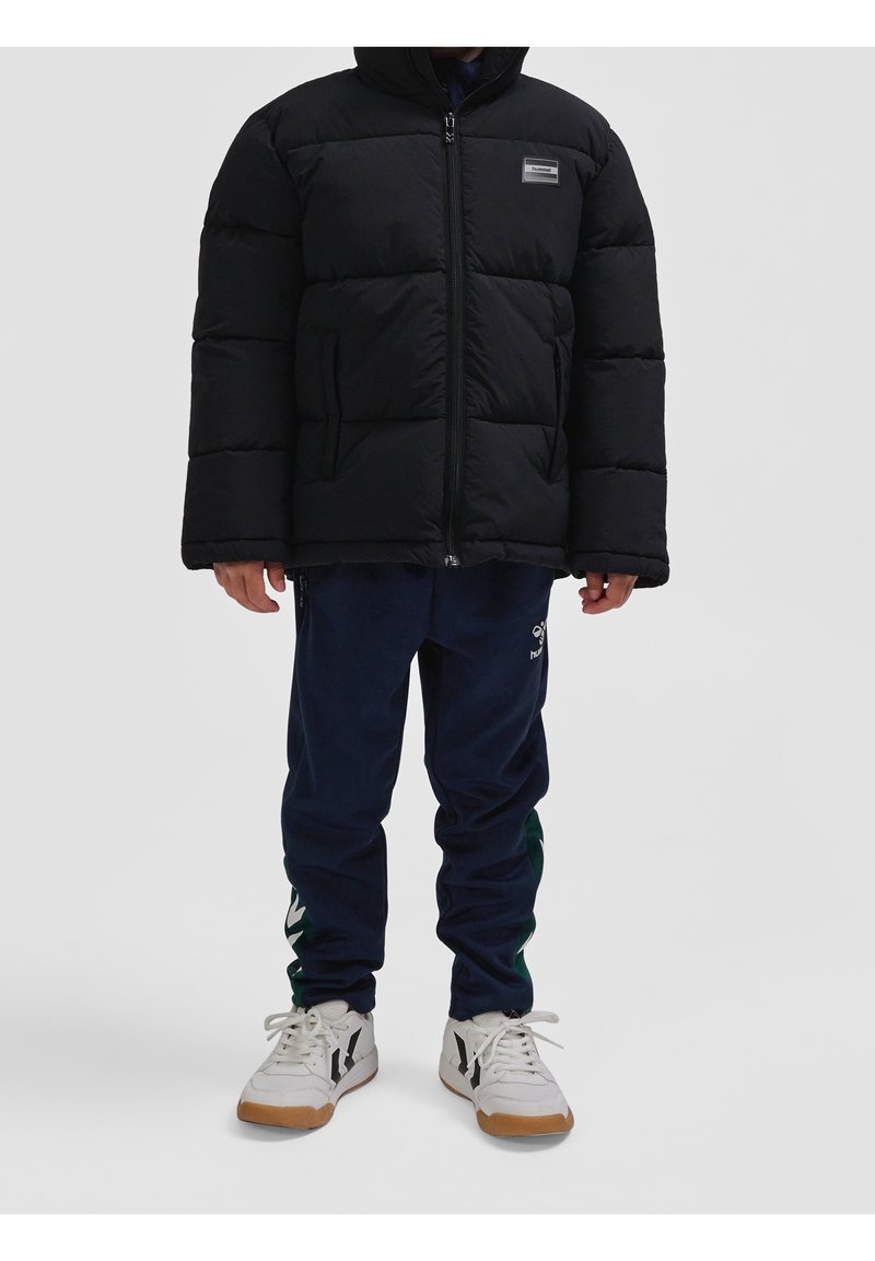 Personne portant une doudoune noire, un pantalon de jogging bleu marine avec des logos et des baskets blanches à semelles en gomme sur un fond uni.
