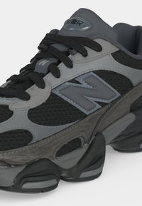 Крупен план на черен и сив маратонка New Balance с текстуриран мрежест материал, дебела подметка и черни връзки.