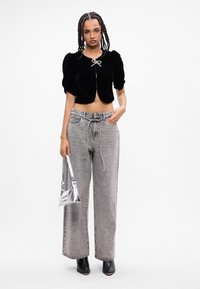 Giacca cropped in velluto nero con maniche a sbuffo e chiusura anteriore a fiocco, abbinata a jeans larghi grigi chiari e una borsa metallica argento.