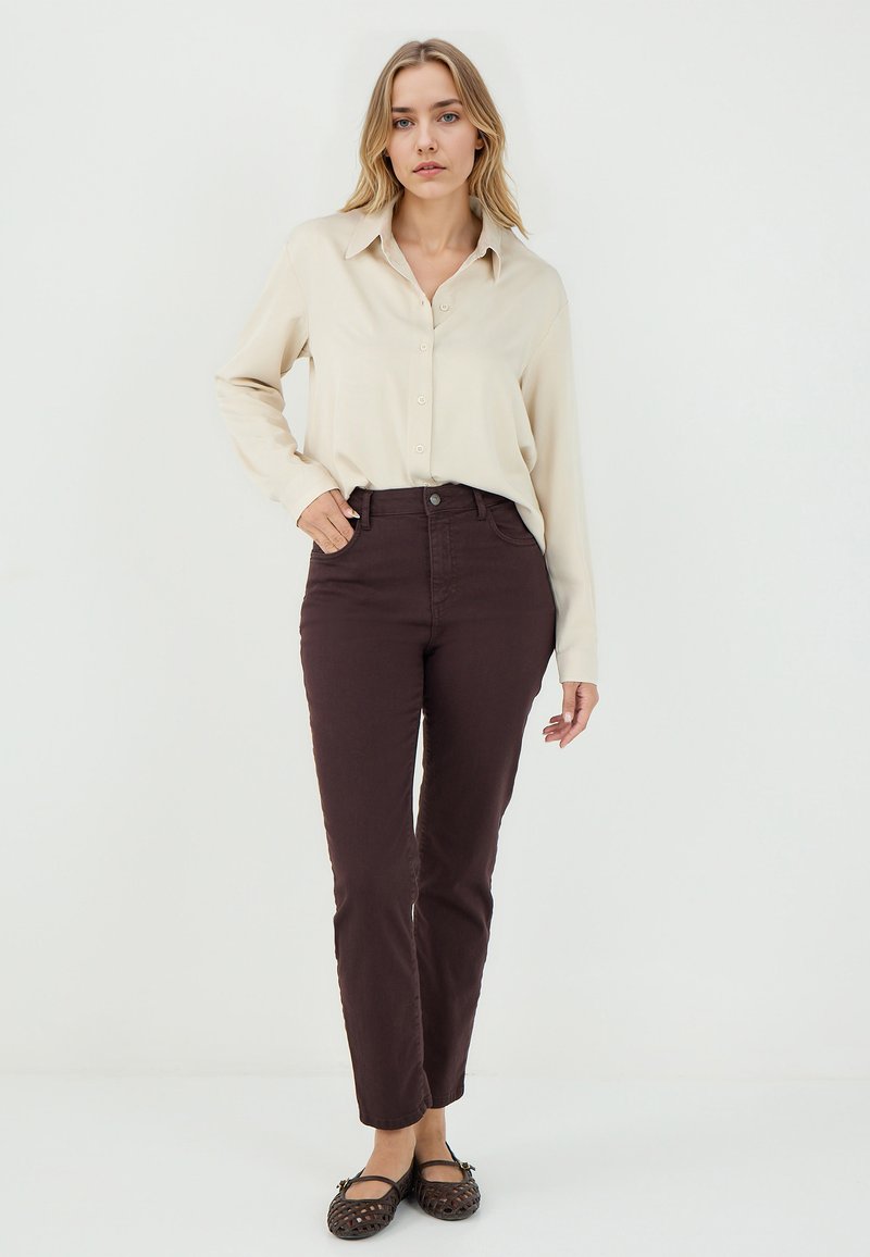 Camicia button-up di un beige chiaro abbinata a pantaloni fitted marrone scuro. La camicia ha maniche lunghe e colletto; i pantaloni sono dritti.