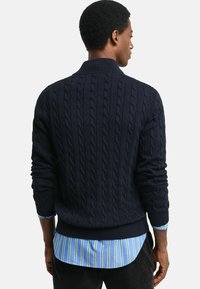 Pull en maille torsadée bleu marine avec des poignets et un ourlet côtelés, muni d'un col montant. Superposé sur une chemise rayée bleu clair.