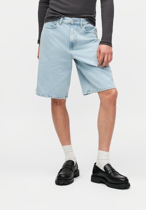 Mężczyzna ubrany w jasnoniebieskie krótkie spodenki jeansowe, czarną koszulę z długim rękawem, białe skarpetki i czarne skórzane mokasyny na jednolitym tle.