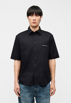 Muž v čiernej košeli s krátkym rukávom zapínanej na gombíky s logom Armani Exchange na hrudnom vrecku a v modrých džínsoch, stojaci na jednoduchom pozadí.