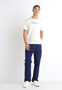 Balts kokvilnas T-krekls ar "Pepe Jeans" logotipu, kombinēts ar ziliem džinsiem. Modelis valkā baltas tenisa kurpes; ikdienišķs piegriezums un viegla tekstūra.