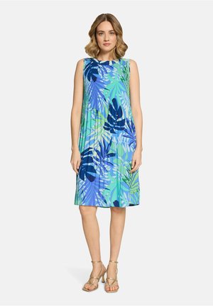 Kobieta stojąca w bezrękawowej sukience sięgającej do kolan, z niebiesko-zielonym tropikalnym wzorem liści, oraz w złotych sandałach na obcasie z paskami.