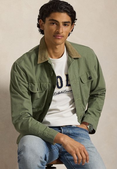 Νέος άνδρας με σγουρά σκούρα μαλλιά, φορώντας πράσινο μπουφάν πάνω από λευκό μπλουζάκι με σχέδιο και μπλε τζιν, καθισμένος με το χέρι ακουμπισμένο στο γόνατο.