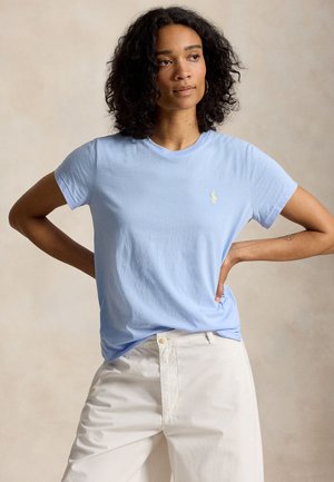 Γυναίκα με σγουρά μαλλιά, που φοράει ένα ανοιχτά μπλε κοντομάνικο μπλουζάκι και λευκό παντελόνι, στέκεται με τα χέρια στη μέση μπροστά σε ουδέτερο φόντο.