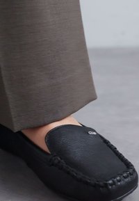 Mustat nahkakengät, joissa on sileä pinta, koristeellinen tikkaus ja etuosassa korostettu logo, yhdistettynä khaki-housuihin.