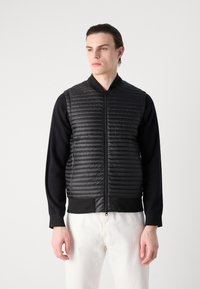 Μαύρο γιλέκο πούφ με οριζόντια ραφή, φερμουάρ μπροστά και ελαστικά τελειώματα. Φορεμένο πάνω από μαύρο μπλουζάκι με μακριά μανίκια, σε συνδυασμό με λευκά παντελόνια.