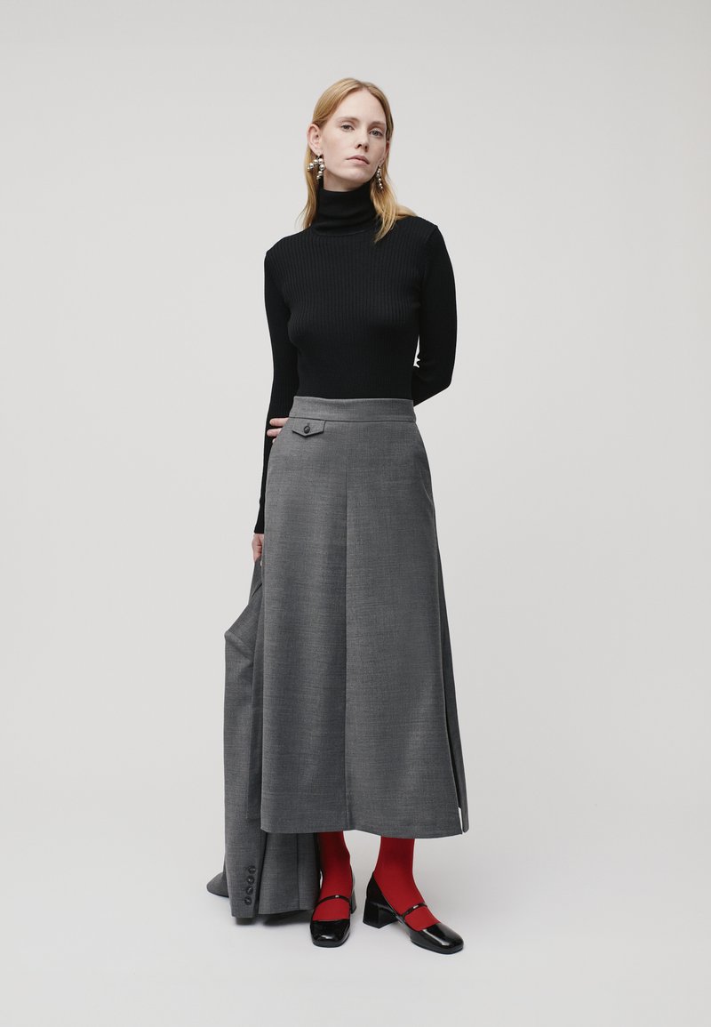 Gonna grigia plissettata con dettaglio a bottoni, abbinata a un dolcevita nero a coste e collant rossi. Scarpe scure completano l'outfit.