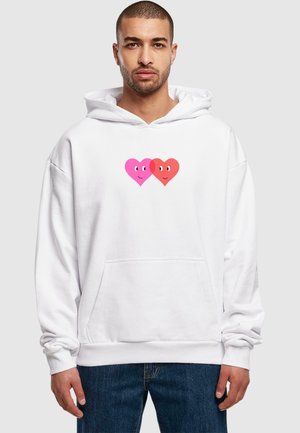 Hvid hættetrøje med en frontlomme, prydet med to tegneserie hjerte- designs i pink og rød. Stoffet er blødt og har en løs pasform.