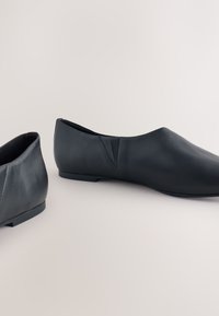Paire de chaussures plates noires en cuir à enfiler avec petits talons et simple couture sur fond clair.