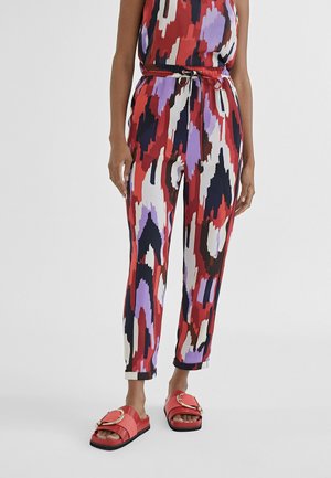 Mujer con pantalones de cintura alta y top con un patrón abstracto en rojo, negro, púrpura y blanco, atado con un cinturón rojo y sandalias rojas deslizantes.