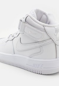 Bijele visokotopne tenisice sa glatkom kožom i mrežastim detaljima, s vezanjem sprijeda, remenom oko gležnja i teksturiranom gumenom potplatom. Nike brendiranje prisutno.