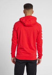 Felpa con cappuccio rossa realizzata in tessuto morbido, caratterizzata da una tasca a marsupio e bande bianche lungo le maniche. Design slim fit.