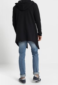 Veste longue à capuche noire en tissu doux, avec un ourlet arrondi. Portée sur un jean bleu clair et des baskets grises avec des accents noirs.