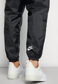 Černé nylonové tepláky s elastickými manžetami, zdobené bílým logem Nike. Nošeny s bílými teniskami Nike Air, které mají texturovanou podrážku.