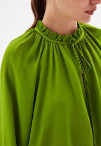 Blusa verde con un colletto arricciato e chiusura a fiocco, caratterizzata da un tessuto fluido e una consistenza morbida, mostrata in un primo piano.