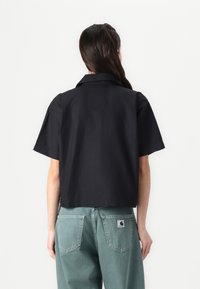 Camisa preta curta com colarinho e mangas curtas, combinada com calças largas de tom verde claro apresentando um remendo no bolso de trás.