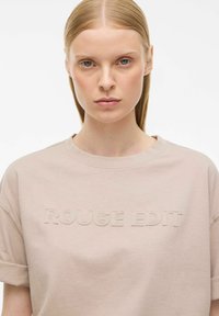 Beżowy t-shirt bawełniany z krótkimi rękawami i okrągłym dekoltem. Zawiera podniesiony napis "ROUGE EDIT" w tym samym kolorze, tworząc subtelną cechę designu.