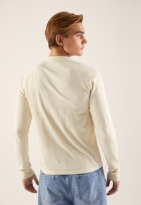 Kremasta majica z dolgimi rokavi z mehko teksturo, raglanskimi rokavi in oprijetimi manšetami, v kombinaciji s svetlo modrimi kavbojkami. Enostaven in nevtralen dizajn.