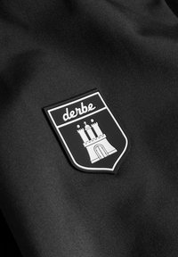 Černá tkanina s vyvýšenou monochromatickou nášivkou zobrazující erb hradu a slovo "derbe" nad ním, lemovaná bílou barvou.