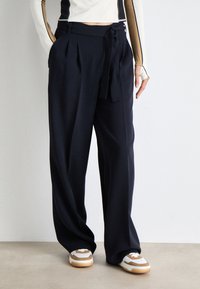 Pantaloni a gamba larga blu navy in un tessuto liscio, caratterizzati da un'alta vita, pieghe frontali e un dettaglio con cintura a fianco. Indossati con sneakers bianche e marroni.