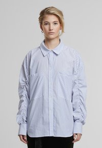Camicia a righe blu chiaro e bianca in tessuto, con colletto, chiusura a bottoni e due grandi tasche frontali.