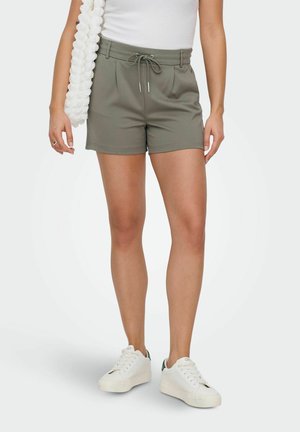 Kobieta nosząca oliwkowo-zielone szorty ze sznurkiem, białe trampki i biały top, trzymająca białą, teksturowaną torbę na ramieniu.