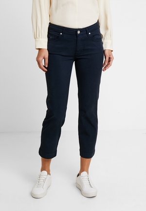 Vrouw draagt donkerblauwe cropped broek, crème blouse met lange mouwen en witte sneakers, staand voor een effen achtergrond.