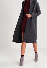 Lungo cappotto grigio realizzato in tessuto morbido, con colletto a scialle e due tasche frontali, indossato sopra un vestito borgogna e stivali alla caviglia grigi.