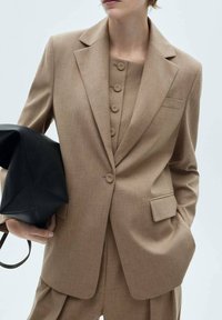 Personne portant un blazer beige boutonné et un pantalon assorti, tenant sous le bras un grand sac en cuir noir texturé.