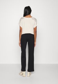 Jasnobeżowa bluzka z koronkowymi detalami na ramionach, zestawiona z czarnymi spodniami i kremowymi butami na klocku, widziana od tyłu.