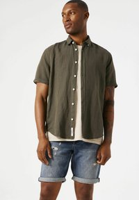 Camisa de manga corta color verde oliva con botones, combinada con shorts de denim azul desgastados. El conjunto presenta un ajuste casual y relajado.