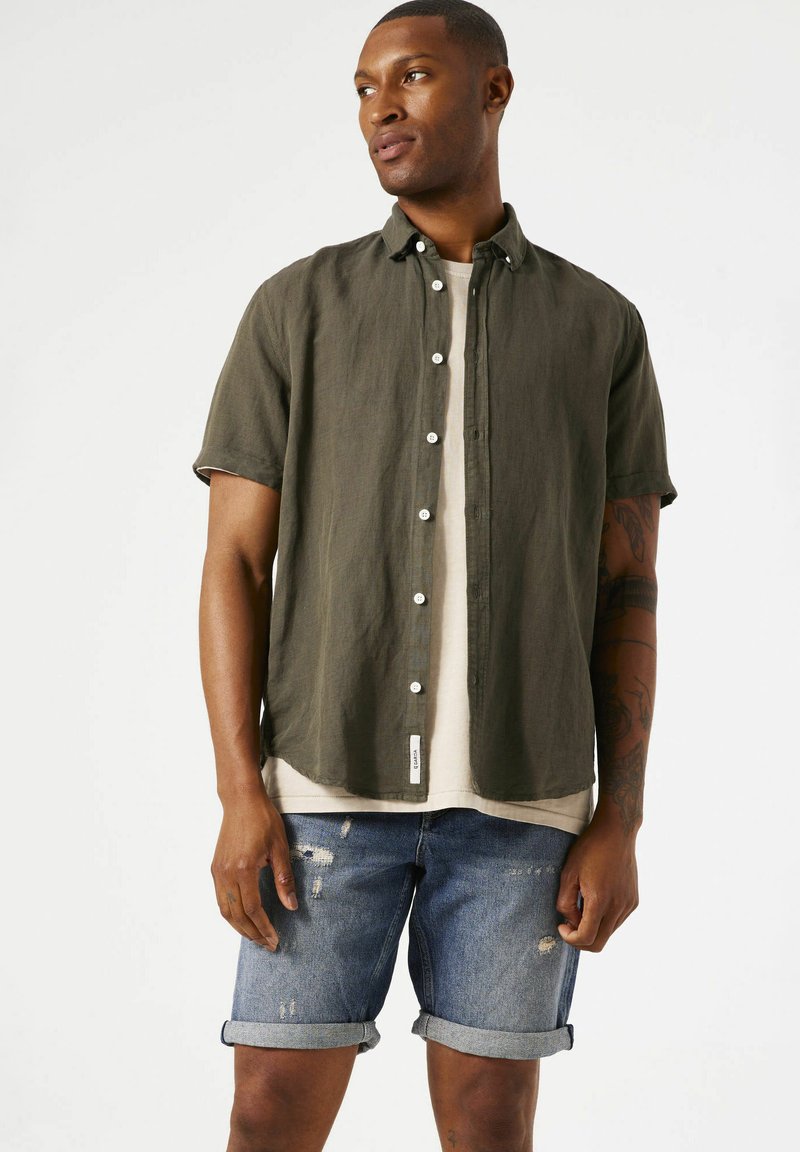 Camisa de manga corta color verde oliva con botones, combinada con shorts de denim azul desgastados. El conjunto presenta un ajuste casual y relajado.
